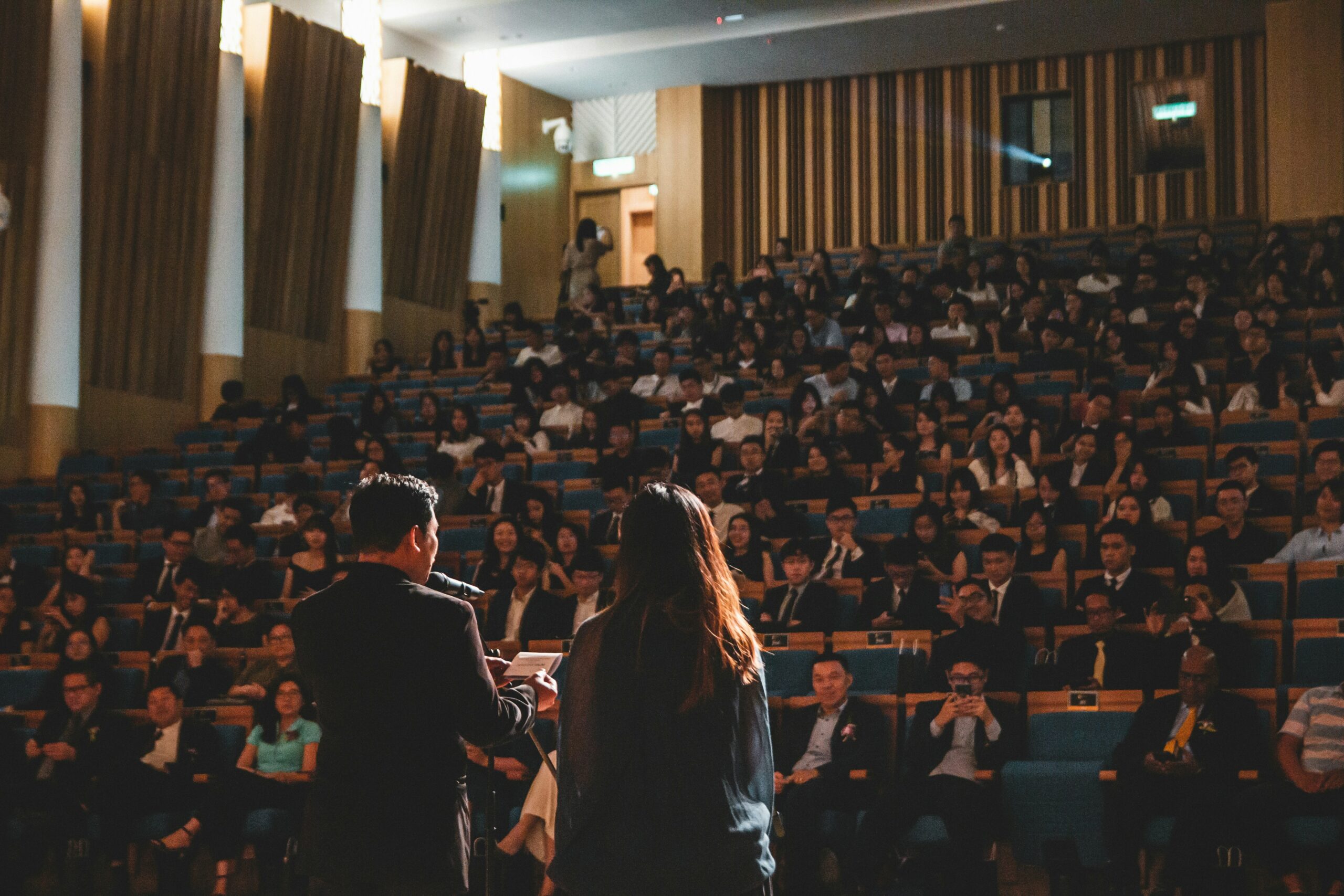 Due persone parlano a un pubblico numeroso in una sala conferenze. Una folla di partecipanti è seduta nelle file di sedili, ascoltando attentamente i relatori sul palco. Agenzia di interpretariato