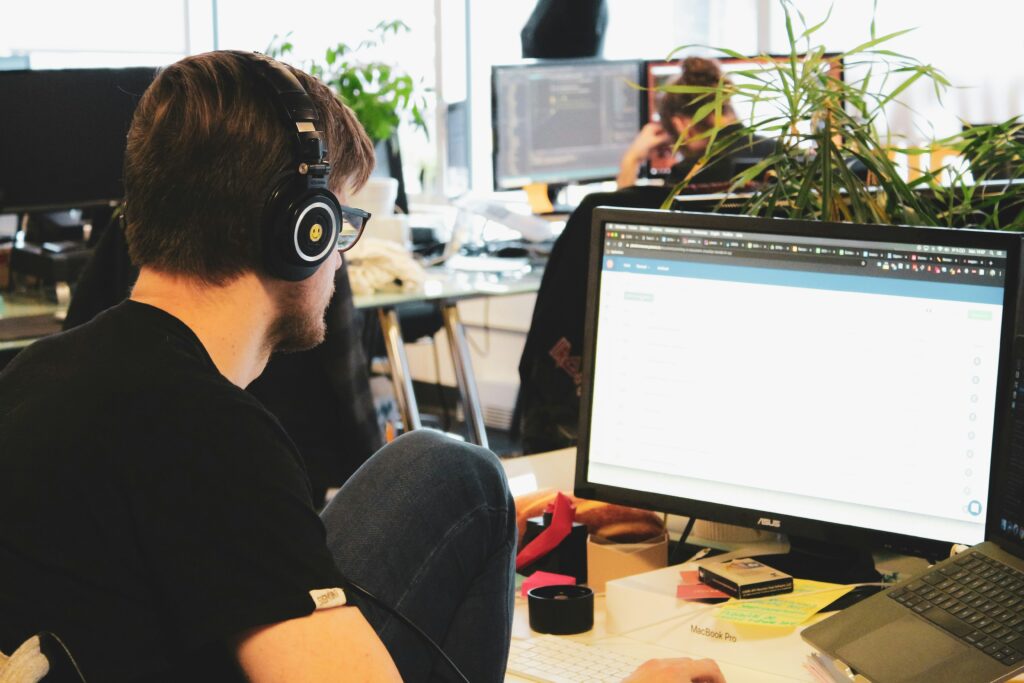 Un uomo con le cuffie lavora in un ufficio aziendale al computer in un ufficio moderno. Sullo sfondo, altri colleghi sono impegnati in attività simili, con vari monitor e piante che decorano lo spazio lavorativo di un'agenzia di interpretariato